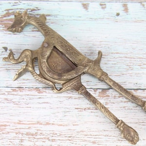 Puede incluir: Cascanueces de latón ornamentado con cabeza de dragón y mangos detallados. Esta herramienta antigua, con mecanismo de palanca, se utiliza para romper nueces. El latón presenta una pátina cálida y envejecida.