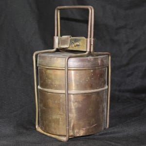 Könnte beinhalten: Vintage zweistufige Lunchbox aus Messing mit Griff. Der Metallbehälter hat einen rechteckigen Boden und einen Klappdeckel. Der Griff ist oben und an den Seiten befestigt. Das Messing hat ein angelaufenes Aussehen.