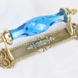 May include: An antique brass door pull with a faceted, light blue glass handle. The rectangular base features ornate detailing and a rope-like border. The hardware is designed for a cabinet or drawer.