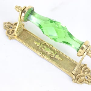 Pode incluir: Um puxador de porta com uma pega de vidro facetado verde brilhante montada numa placa traseira retangular de latão. A placa de latão apresenta um padrão em relevo ornamentado com volutas decorativas em ambas as extremidades e um design central.