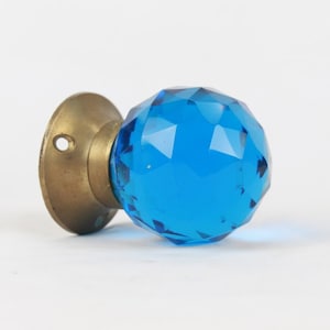 Puede incluir: Un pomo de puerta de cristal azul con base de latón. El pomo es facetado y tiene un acabado brillante.