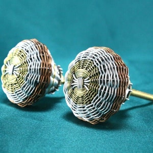 May include: Two decorative cabinet knobs made of woven wire in silver, gold, and copper tones. The knobs have a round shape and a threaded stem.