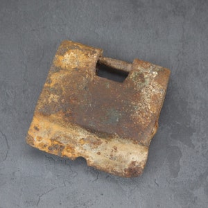 Puede incluir: Un candado de metal oxidado con forma cuadrada y un ojo de cerradura. El candado está desgastado y muestra signos de antigüedad.