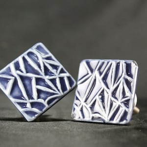 Op de afbeelding: Twee vierkante keramische kastknoppen met een wit geometrisch patroon op een donkerblauwe achtergrond.