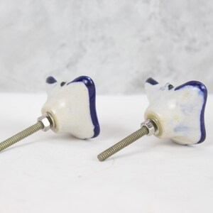 May include: Two white ceramic cabinet knobs with blue trim. The knobs are shaped like a flower with four petals. Each knob has a silver screw.