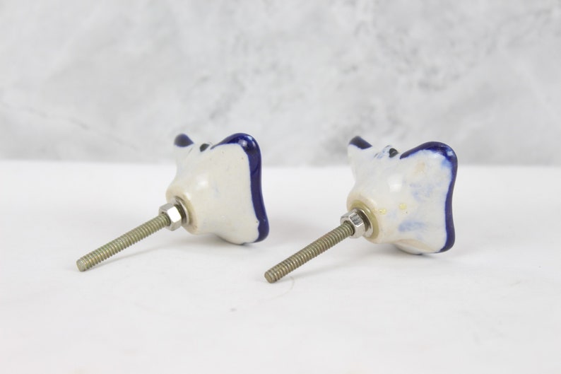 May include: Two white ceramic cabinet knobs with blue trim. The knobs are shaped like a flower with four petals. Each knob has a silver screw.