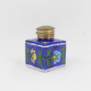 May include: A small blue ceramic inkwell with a brass lid. The inkwell is decorated with a floral design in yellow, green, and blue.