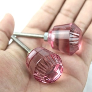 Puede incluir: Dos pomos de armario de cristal rosa con un diseño facetado y tornillos plateados.