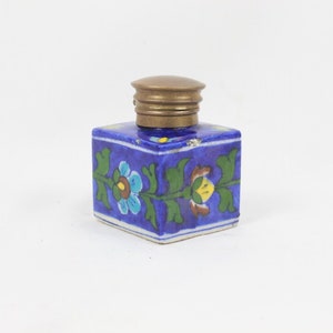 May include: A small blue ceramic inkwell with a brass lid. The inkwell is decorated with a floral pattern in shades of green, yellow, and blue.