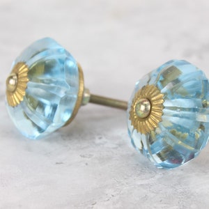 Peut inclure: Deux boutons de cabinet en verre bleu clair avec des accents en métal doré. Les boutons ont un design facetté et sont fixés à des vis en métal.