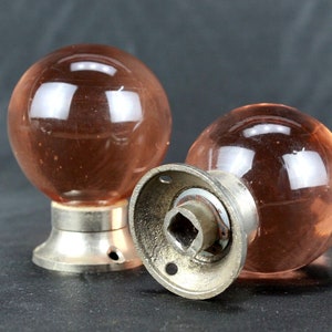 Puede incluir: Dos pomos de puerta de vidrio rosa con bases de metal plateado. Los pomos son redondos y tienen un acabado liso y brillante.