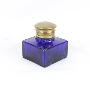 May include: A small, square, cobalt blue glass inkwell with a brass lid. The lid has a slightly domed top and a decorative band around the base.
