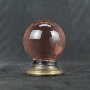 Peut inclure: Une sphère en verre rose avec une base en laiton. La sphère est transparente et a une légère teinte rose. La base est ronde et a une bande noire autour du haut.