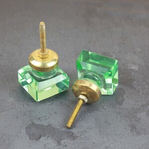 Könnte beinhalten: Zwei grüne Glas-Schrankknöpfe mit goldenen Metallsockeln und Schrauben. Die Knöpfe sind quadratisch mit abgeschrägten Kanten.