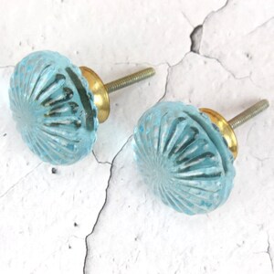 Op de afbeelding: Twee blauwe glazen kastknoppen met een gouden schroef. De knoppen hebben een decoratief, zonnestralen-achtig patroon.