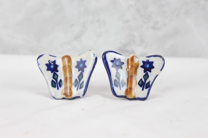May include: A pair of ceramic butterfly drawer pulls with blue and white floral designs. The butterflies have brown centres and green leaves.