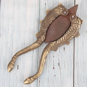 Peut inclure: Casse-noix vintage en laiton avec un motif de poisson. Le casse-noix est doté d'une lame en métal foncé et de poignées ornées. Les poignées et les extrémités en forme de poisson sont en laiton. Sur une surface en bois bleu clair et blanc.