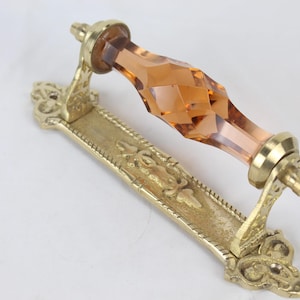 May include: Ornate brass door handle with a faceted amber-colored glass center. The handle features decorative floral and scrollwork details on the base and mounting hardware. A vintage-style hardware piece.
