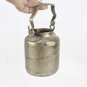 May include: A vintage metal container with a handle. The cylindrical body has horizontal bands and a tarnished finish. The handle is arched and attached with rings. The container is held against a white background.