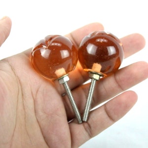 May include: Two amber glass cabinet knobs with silver screws. The knobs have a rounded shape with a slightly textured surface.