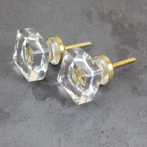 Op de afbeelding: Twee heldere glazen kastknoppen met gouden metalen bases en schroeven. De knoppen zijn vierkant met afgeschuinde randen.