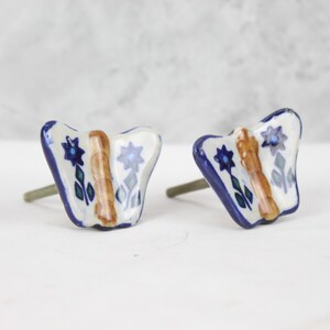 May include: Two ceramic butterfly drawer knobs with a white background, blue and green floral accents, and a brown stripe down the centre.