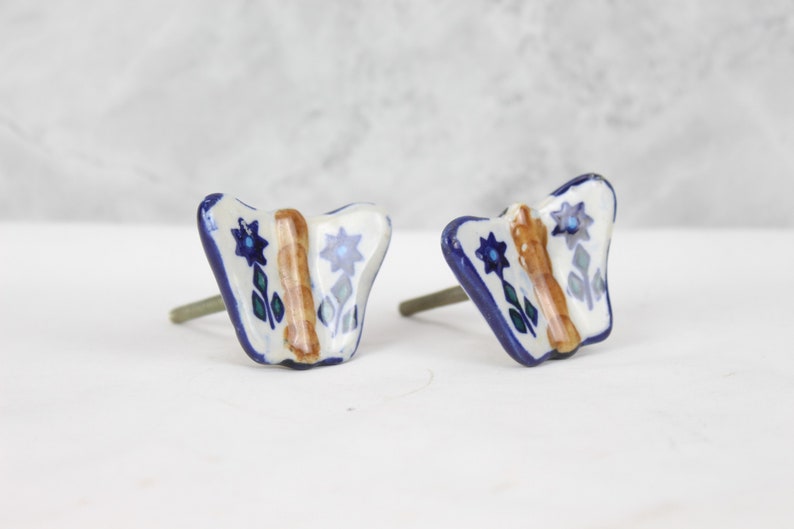 May include: Two ceramic butterfly drawer knobs with a white background, blue and green floral accents, and a brown stripe down the centre.