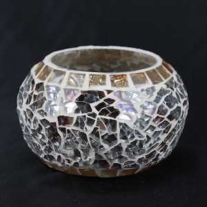 Puede incluir: Un portavelas redondo de mosaico blanco con un borde dorado. El portavelas está hecho de pequeñas piezas de vidrio iridiscente.