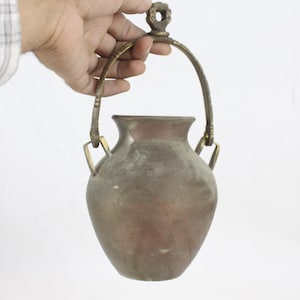 Puede incluir: Un jarrón de metal de color bronce vintage con asa. El jarrón tiene un cuerpo redondeado y un cuello estrecho. El asa es arqueada y está sujeta al jarrón con soportes decorativos. El jarrón está sostenido por una mano.
