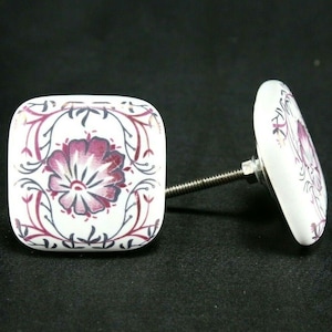 May include: Two white ceramic square cabinet knobs with a floral design in shades of pink and gray. The knobs have a silver screw.
