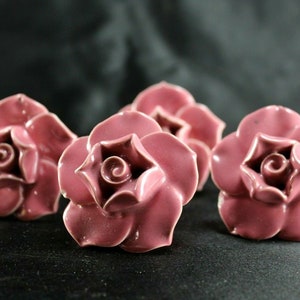 May include: Four ceramic rose-shaped drawer pulls in a dusty rose color. The roses have a detailed, three-dimensional design.