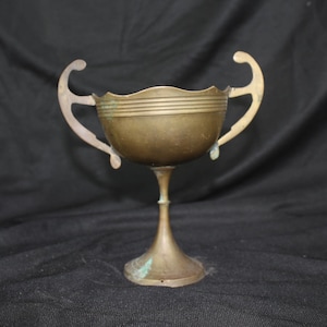 May include: A vintage brass trophy cup with two curved handles. The cup has a round bowl with horizontal lines and a scalloped rim. The base is a flared pedestal. The brass has a patina, with some green oxidation visible.