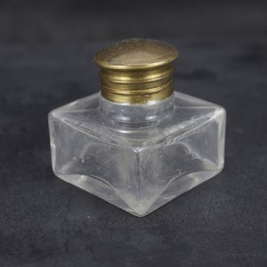 Può includere: Un calamaio di vetro trasparente con un coperchio in ottone. Il calamaio è di forma quadrata e ha una parte superiore leggermente arrotondata. Il coperchio è un semplice disco piatto con un bordo leggermente rialzato.