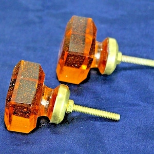 Könnte beinhalten: Zwei Schrankknöpfe aus bernsteinfarbenem Glas mit Messingsockeln und Schrauben. Die Knöpfe sind achteckig und haben eine strukturierte Oberfläche.
