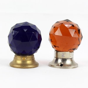Op de afbeelding: Twee glazen deurknoppen met gefacetteerde oppervlakken. De ene knop is blauw en de andere is oranje. De knoppen zijn gemonteerd op metalen bases.