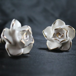 May include: Two white ceramic rose-shaped drawer pulls with gold accents. The pulls have a screw on the back for attaching to a drawer.