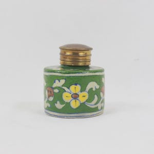 May include: A small cylindrical ceramic inkwell featuring a vibrant green base color with a hand-painted floral pattern in yellow, white, and brown. The container is topped with a metallic brass-colored screw-on lid with horizontal ridges.
