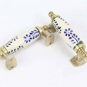 以下が含まれることがあります： 2つの装飾的なキャビネットハンドル。各ハンドルは、青い花の模様と真鍮の金具が付いたクリーム色のセラミックグリップが特徴です。これらのハンドルは、家具の修復や新しいキャビネットにエレガントさを加えるのに適しています。