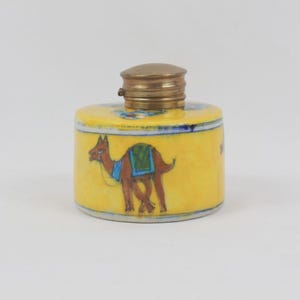 May include: A cylindrical yellow ceramic snuff bottle featuring a hand-painted brown camel with a blue and green saddle. The bottle is topped with a metallic brass-colored cap that has a ridged texture and a small hinge on the side.