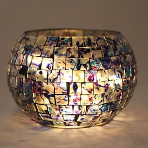 Puede incluir: Un portavelas redondo de vidrio con un diseño de mosaico en tonos azules, morados y blancos. El portavelas está iluminado desde el interior, creando un brillo cálido.