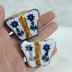 May include: Two ceramic butterfly-shaped ornaments with blue and white floral designs. The butterflies have brown centres and are painted in a folk art style.