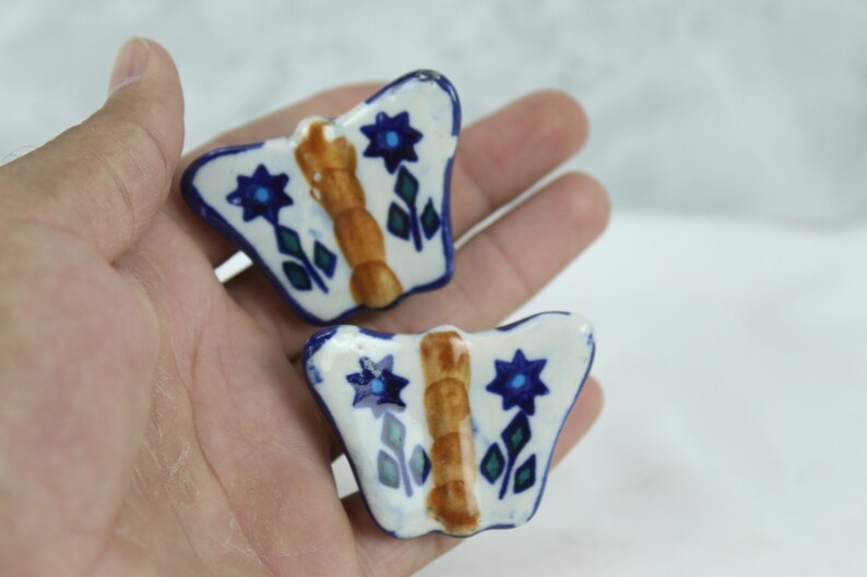 May include: Two ceramic butterfly-shaped ornaments with blue and white floral designs. The butterflies have brown centres and are painted in a folk art style.