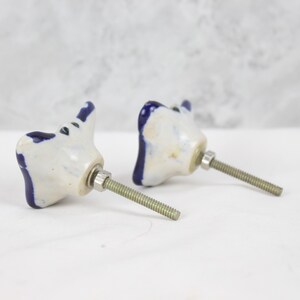 May include: Two white ceramic drawer pulls with blue accents in the shape of a stylised butterfly. Each pull has a silver screw.