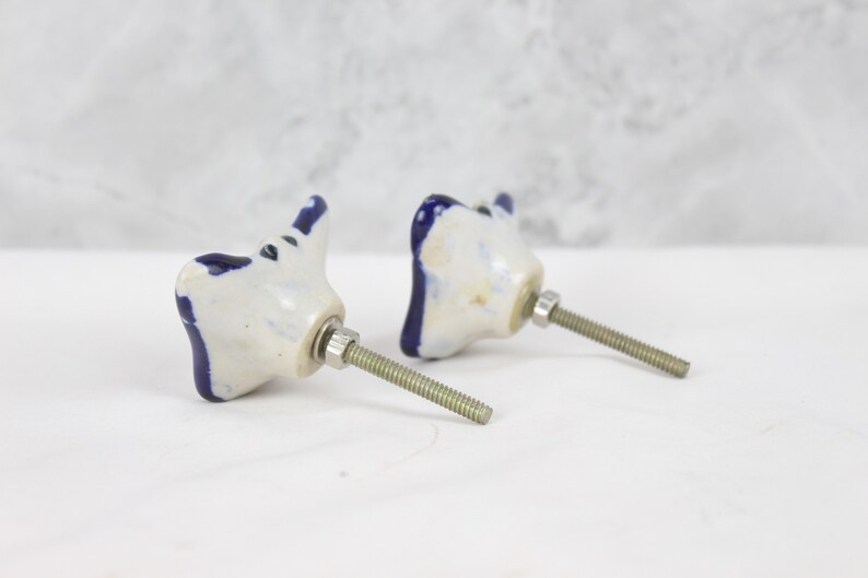May include: Two white ceramic drawer pulls with blue accents in the shape of a stylised butterfly. Each pull has a silver screw.