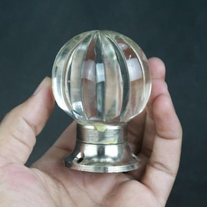 Puede incluir: Un pomo de puerta de cristal transparente y acanalado con una base de metal plateado. El pomo es redondo y tiene un diseño decorativo y vintage. La imagen es un primer plano del pomo sostenido en una mano sobre un fondo oscuro.