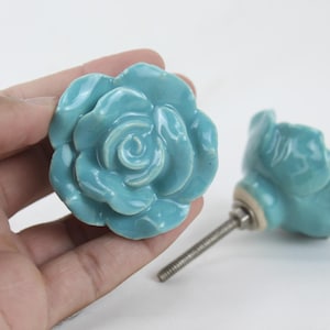May include: Two ceramic rose-shaped drawer pulls in a light blue colour. The pulls have a detailed rose design and are attached to a silver screw.