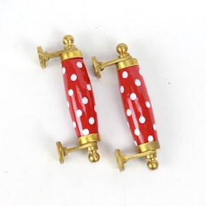 Puede incluir: Dos pomos de armario de cerámica roja con lunares blancos y detalles en metal dorado. Los pomos son de forma ovalada y tienen un pequeño pomo redondo en la parte superior.