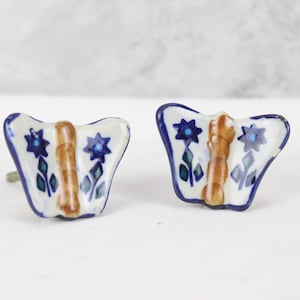 Può includere: Due tiretti per cassetti in ceramica a forma di farfalla con motivi floreali blu e marroni. I tiretti sono bianchi con accenti blu e marroni.