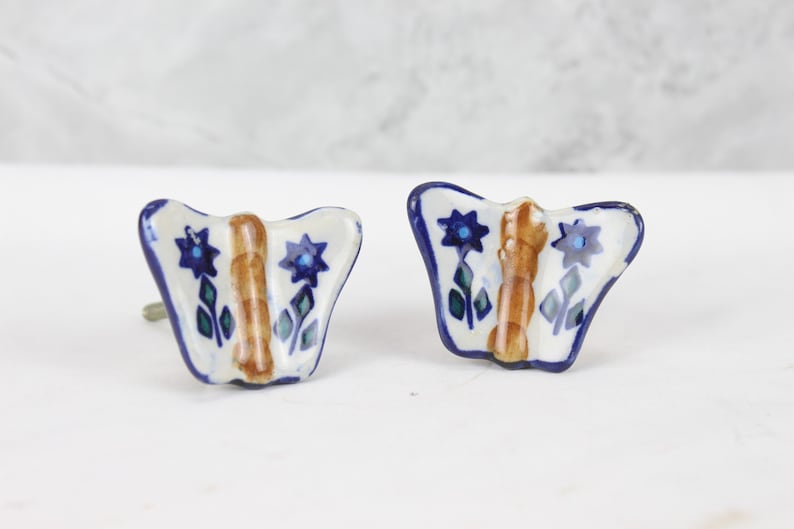 May include: Two ceramic butterfly drawer pulls with blue and brown floral designs. The pulls are white with blue and brown accents.