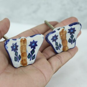 May include: Two ceramic butterfly drawer knobs with blue and green floral designs and a brown centre. The knobs have a silver screw.
